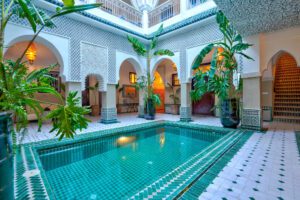 The interior courtyard of a traditional Moroccan riad, a perfect reason to book accommodation in Morocco, featuring an inviting swimming pool with green mosaic tiles, white and green tiled floors, ornate archways, and lush green plants.
