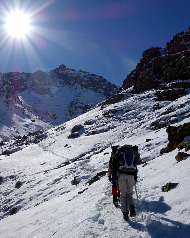 Two mountaineers with backpacks and trekking poles hike up a snowy mountain path under a bright, sunny sky in the High Atlas Mountains of Morocco.