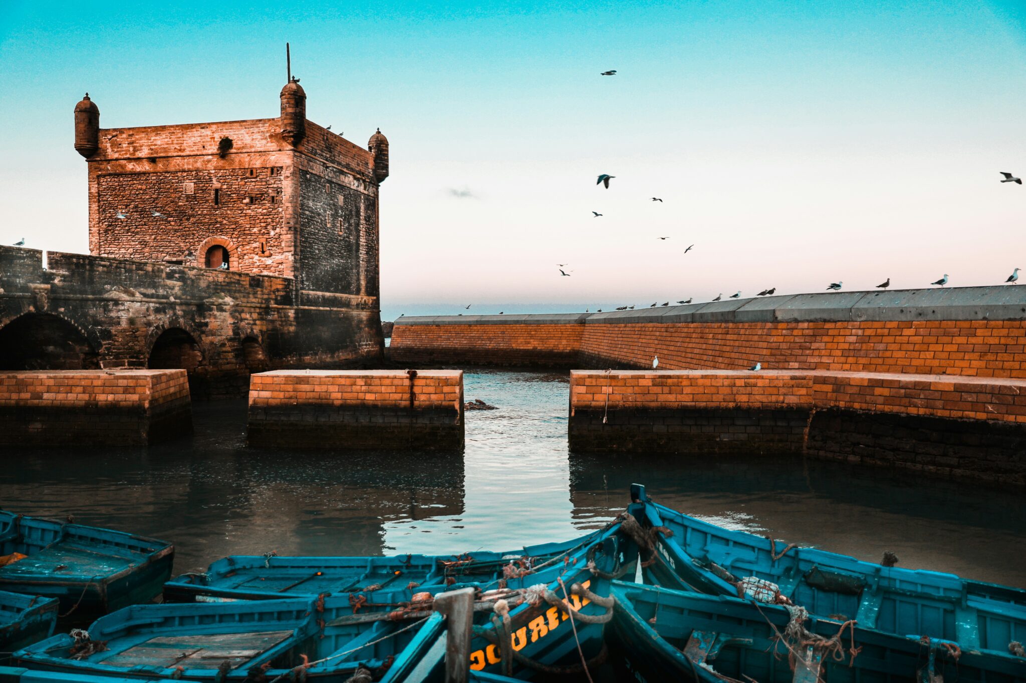 The historic Sqala du Port ramparts in Essaouira, Morocco, with traditional blue fishing boats in the harbor and seagulls flying against a bright blue sky.