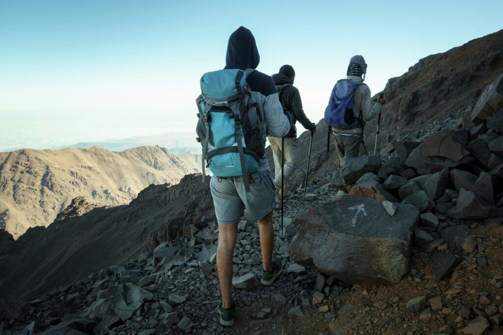 A group of hikers with backpacks and trekking poles navigate a rocky mountain ridge in the High Atlas Mountains, Morocco, overlooking a vast mountain range.